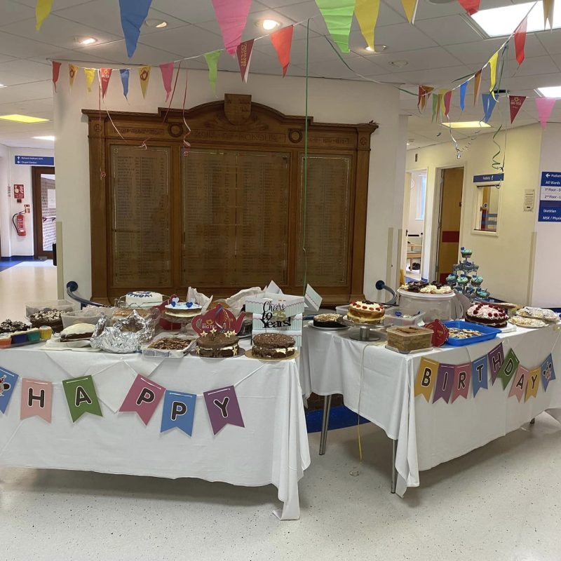 Two trestle tables with white table cloths stand in front of the War Memorial. Bunting spelling Happy Birthday hangs from the tables. The tables are full of colourful cakes, cupcakes, biscuits and treats. Above the tables colourful bunting hangs from the ceiling.