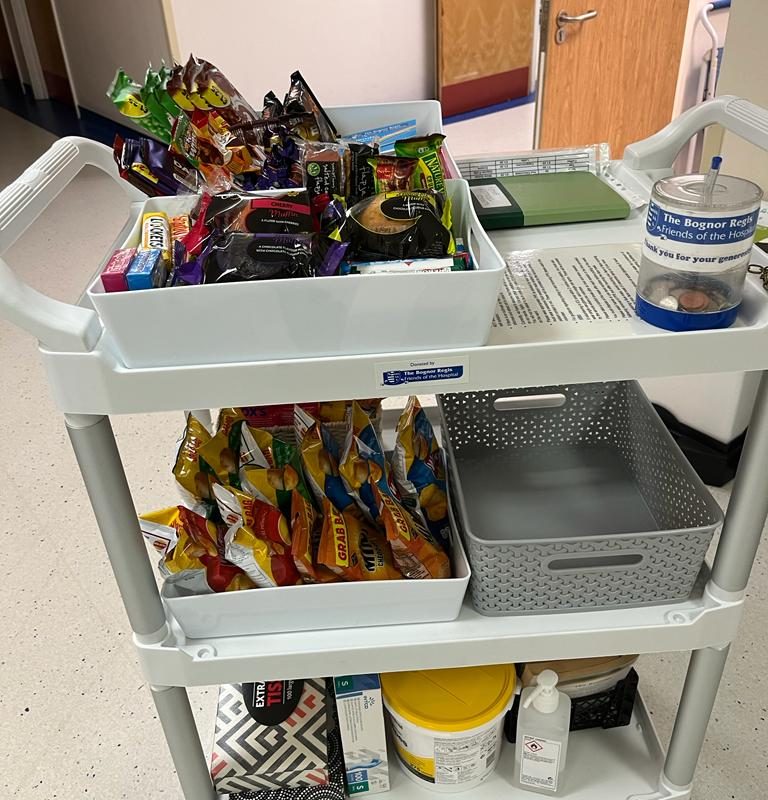 Picture shows the tea bar trolley. Metal legs with three white shelves, stocked with edible and personal care items for sale.