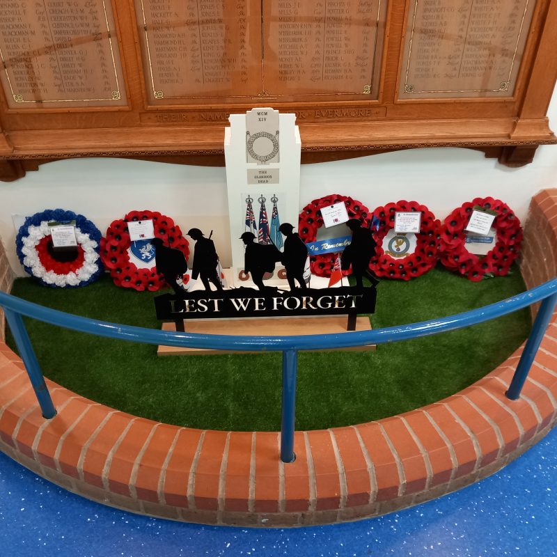 Picture shows the War Memorial at Bognor Hospital, with 5 poppy wreaths and a silhouette sculpture of 5 servicemen saying Lest We Forget, in front of the framed list of names