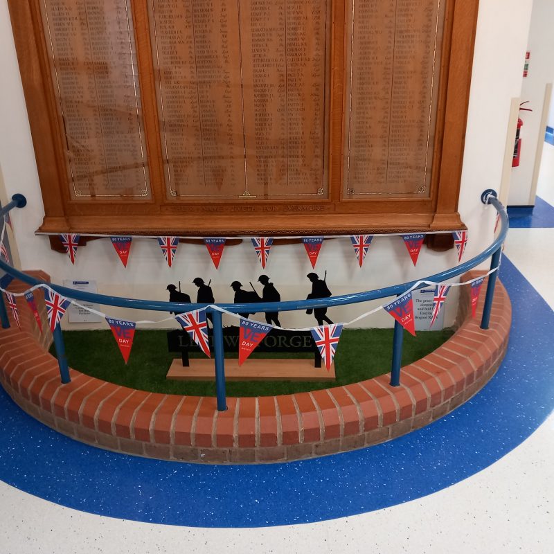 The War Memorial Roll of Honour is pictured with black silhouetted military figures in front, and the Memorial is string with union flag bunting