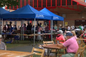Two stalls under blue Friends liveried covers. The stall tables are covered with books for sale. Attendees sit at trestle tables. 