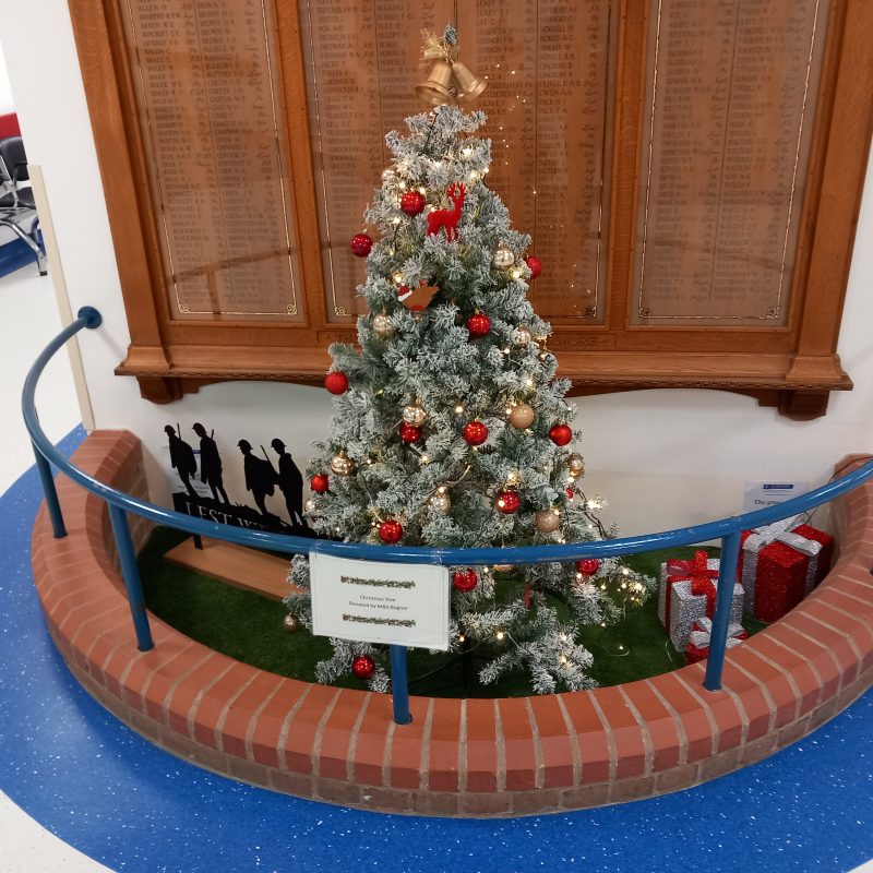 The war memorial in Bognor Hospital is shown with a snowy Christmas tree in front of it with cheerful red and gold baubles, with two gold bells topping the tree. At teh base are decorative presents in red paper with silver bows.