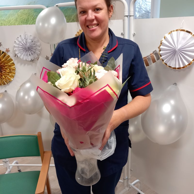 Ward Sister Kathryn Tusker stands in the ward holding a glass vase with white flowers in pink cellophane