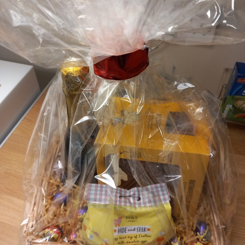 A gift wrapped hamper in a wicker basket with a bottle of bubbly, Easter chocolate and other goodies